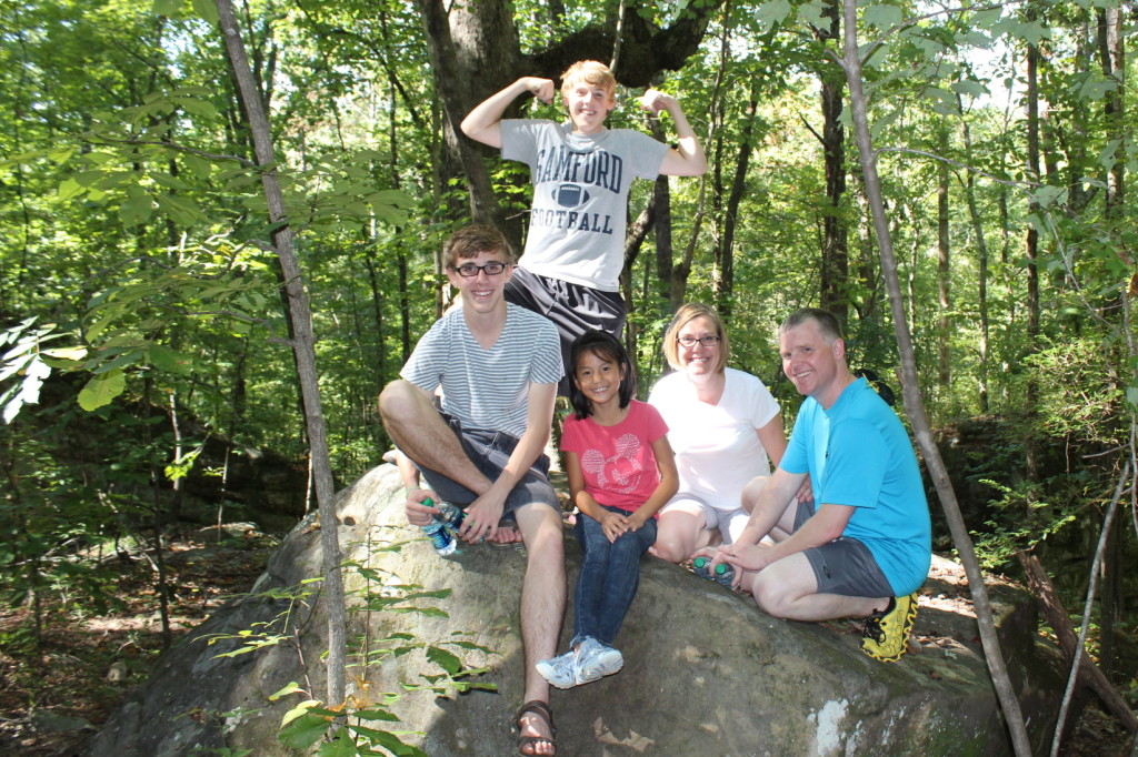 Family smiling on a visit to Moss Rock Preserve, Hoover forest preserve in Alabama. Four sitting on rock and one standing behind 