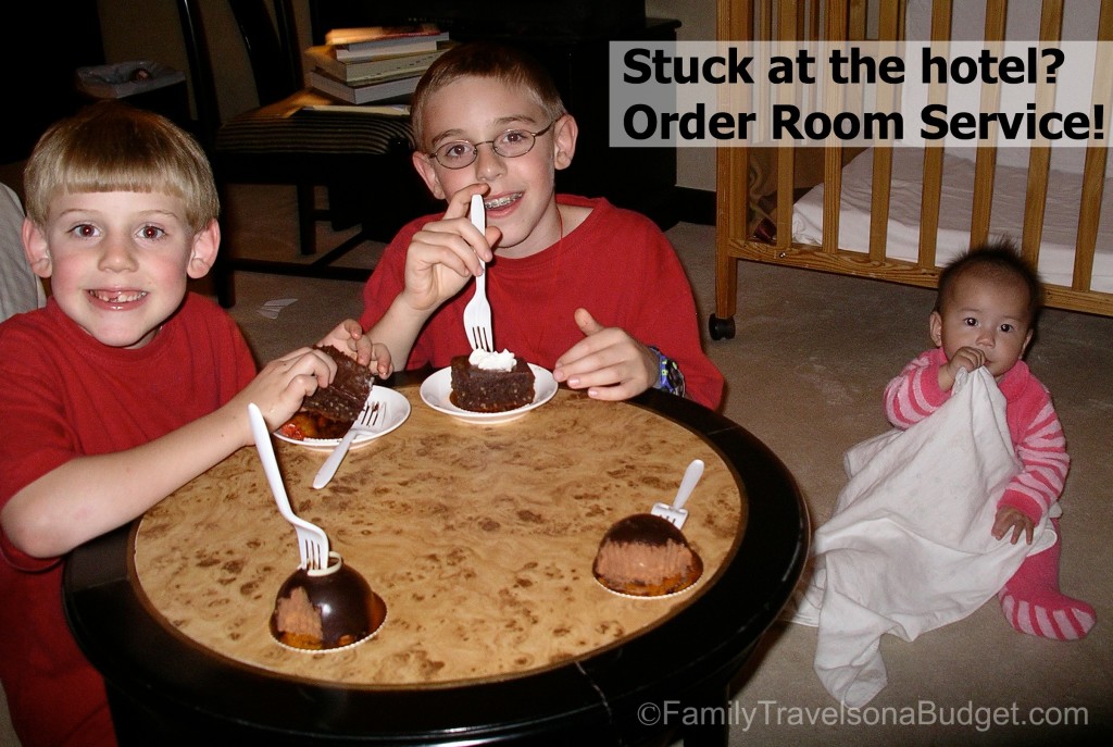 Kids eating chocolate cake at a table in a hotel room with baby sitting on the floor next to them and a crib in the background.