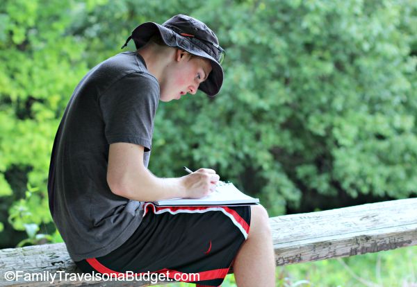 Teenage boy in shorts and hat, sketching while sitting on a railijng at Pere Marquette State Park