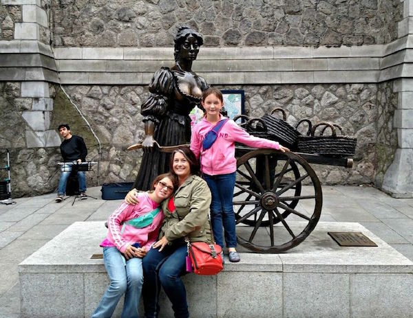 Familiy posing in front of a stature of a woman with a wagon and baskets, with a man playing music in background by a stone wall