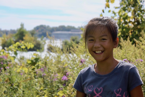 Young girl smiling with wildflowers and the Niagara River in the background at the Niagara Gorge hiking trails.