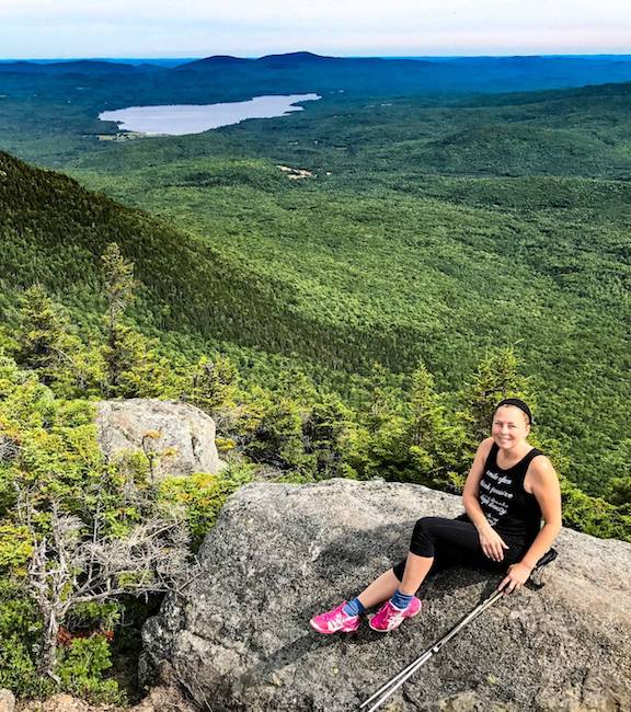 Woman looking up at camera from a rock outcropping at Tumbledown Mountain hiking trails in Maine with green trees below and a lake off in the distance