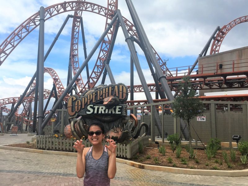 Young teenager in gray top and sunglasses standing in front of the Copperhead Strike roller coaster at Carowinds.