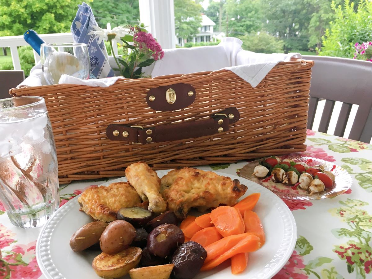 Picnic basket with fried chicken, vegetables, wine and flowers served at a flower covered picnic table.