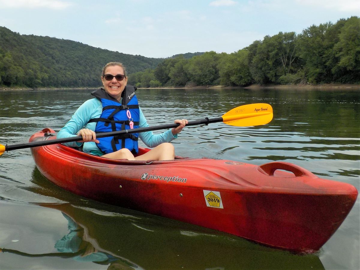 Lady in blue life jacket with sunglasses on a red kayak with a yellow paddle.