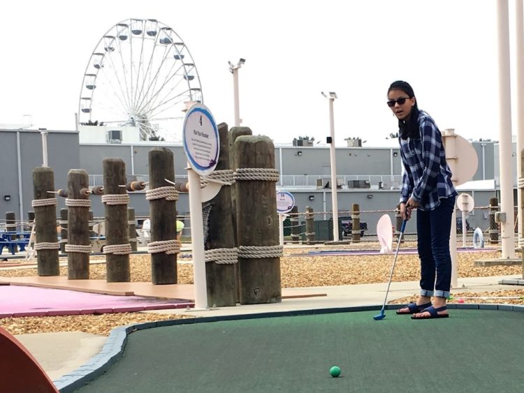 Teen girl in blue plaid on a putt putt course with a ferris wheel in background.