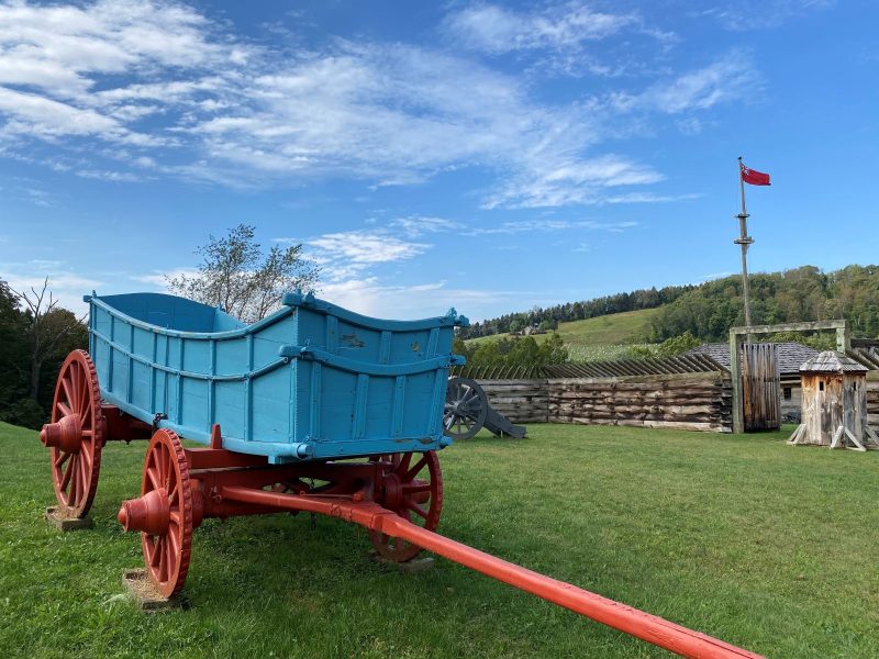 Blue, wooden supply wagon with red wheels showing iron knobs to cross rough terrain with Fort Ligonier, British Flag and Laurel Highlands behind
