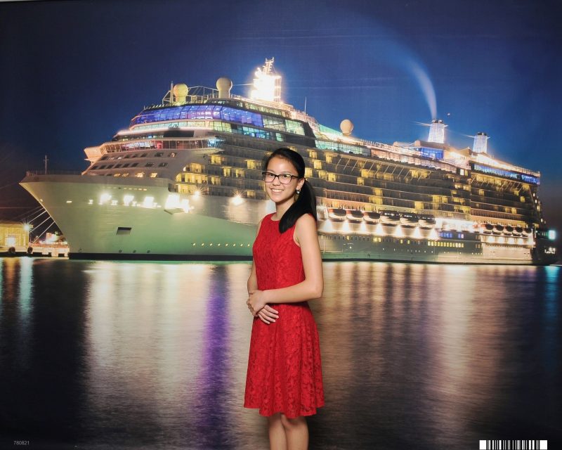 Teen girl in red dress standing in front of a cruise ship at night. Celebrity Equinox is all lit up.