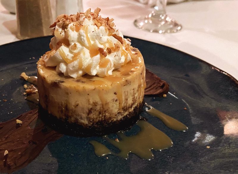 Turtle cheesecake on a blue stoneware plate. Chocolate crust under the cheesecake, with caramel sauce, toasted pecans and whipped cream