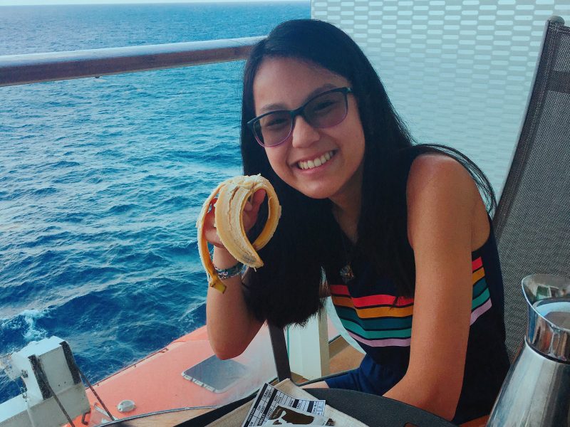 Teen in blue jumper and sunglasses eating banana on ship balcony at sea