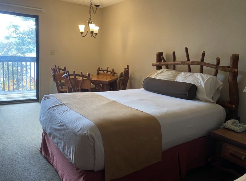 Queen size bed, four chairs and a table with light above and a view of the balcony in a lodge room at Lake Raystown Resort