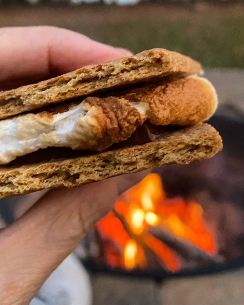 CLose up of a s'more with the toasted marshmallow between two graham crackers with chocolate and a campfire blurred in the background