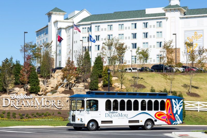 Entrance sign to Dollywood DreamMore Resort with resort building behind and the complimentary theme park shuttle in the foreground. Blue and white Shuttle has butterflies on its side.
