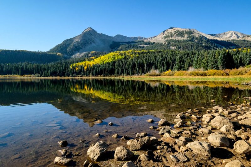 Views at Lost Lake Campground in Colorado, one of the best campgrounds for families in the US. Image shows mountains with pine trees, and clear lake with reflection and rocky bottom.

