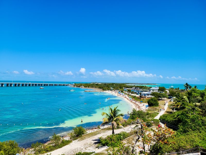 Bahia Honda in the Florida Keys has some of the best camping in Florida for families. Image shows Caribbean blue waters on a crescent beach with a causeway bridge in the distance.