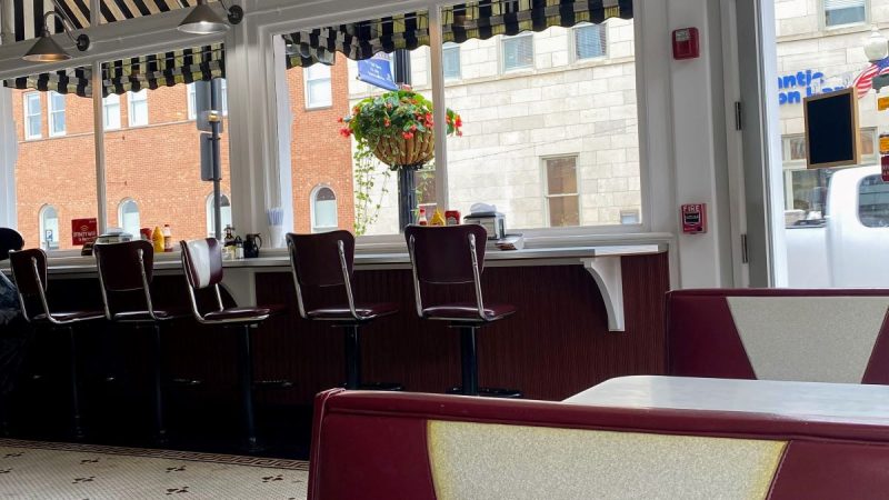 Diner restaurant with red, vinyl bar stools along a window with a black and white stripe awning and baskets of flowers outside the window.