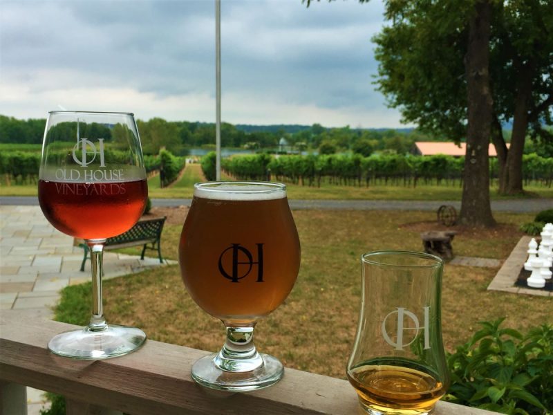 A trio of drinks at Old House Vineyards, with wine, beer and spirits in glasses in the foreground. Behind, the vineyard and giant chess board on the grounds can be seen.