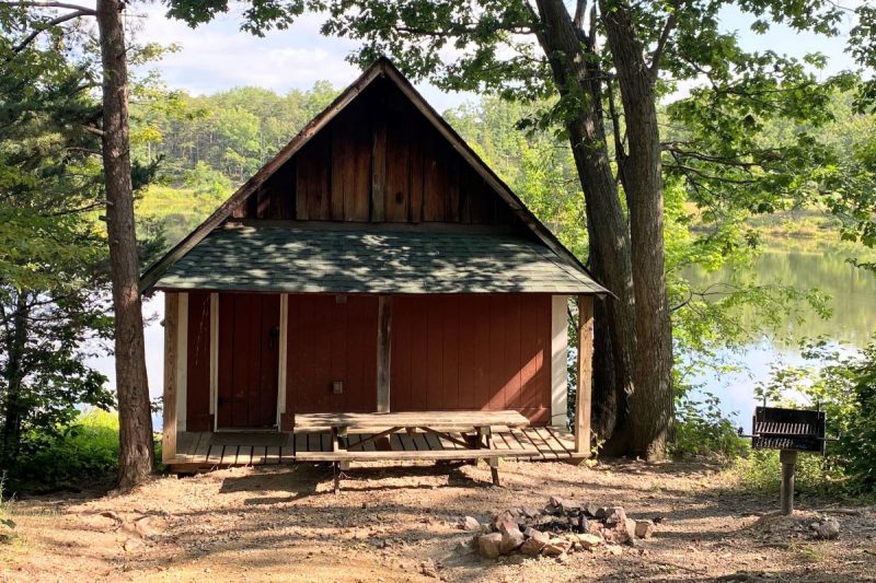 Rustic red cabin with covered front porch, picnic table, fire pit and charcoal grill next to the lake a Cove Camping in the Virginia mountains