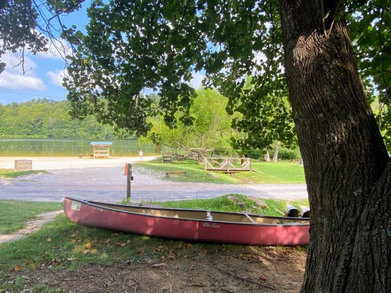 Red Old Town canoe sitting under a tree with beach, swimming lake and hills in the background