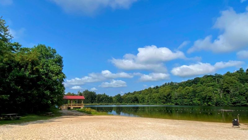 Sandy beach in foreground, with red roof pavilion in background, and trees on the hills beyond. Blue sky with white puffy clouds overhead