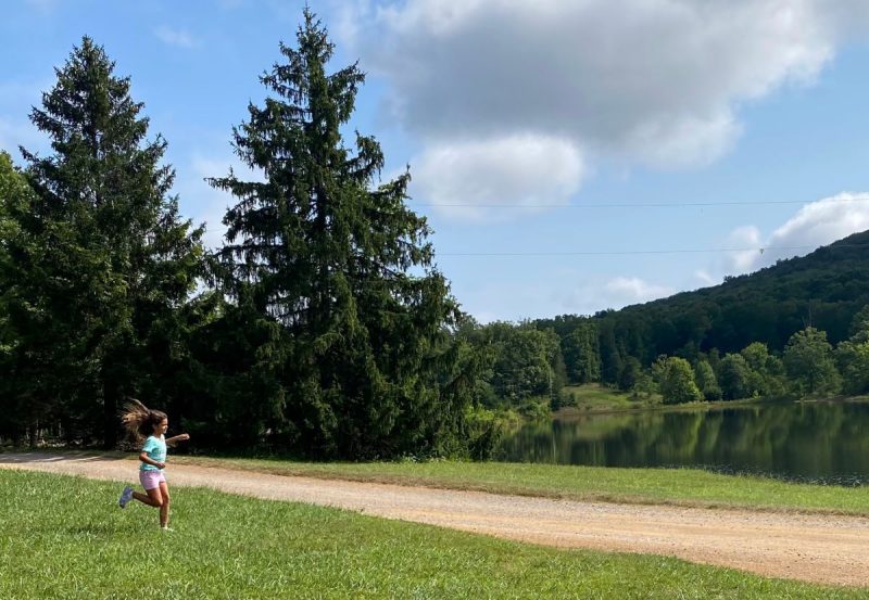 Child running through field at a campground in the Virginia mountains with dirt road, evergreen trees and lake