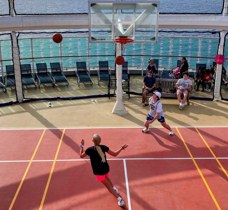 teens on a cruise ship playing basketball on the sports court. Girl with blond ponytail in black shirt and pink shorts. Boy in light colored shirt with white ball cap and blue shorts. Other people in background