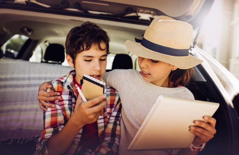 Two kids making car scavenger hunt list sitting in hatchback of a car. Girl has hat and boy has pen and notebook.