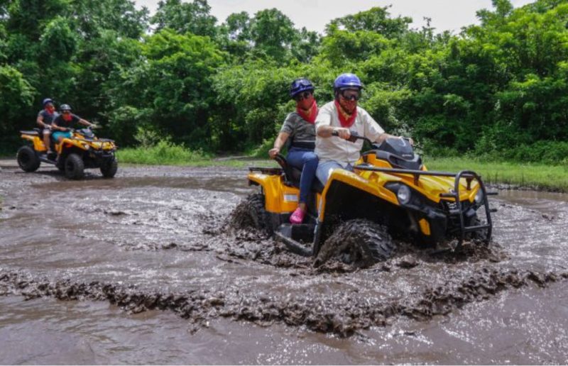 Two yellow ATVs with riders going through a muddy creek in Cozumel Mexico wearing helmets and goggles