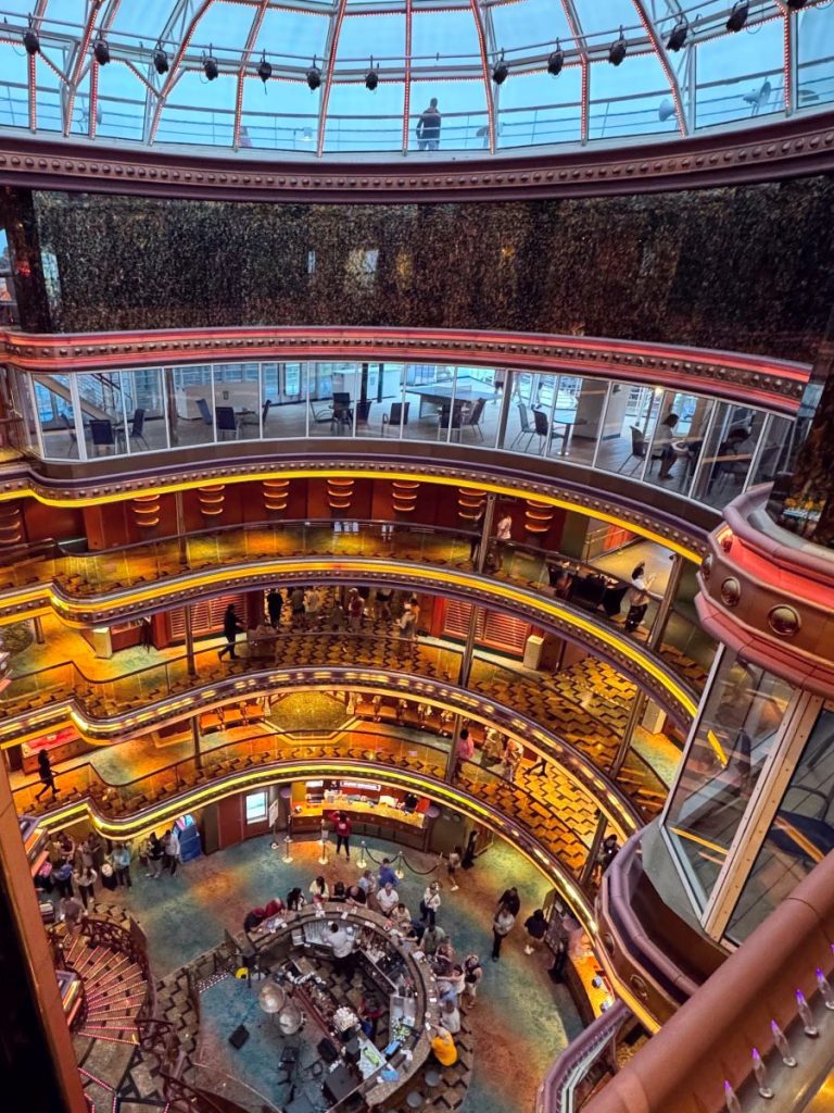 Carnival Elation atrium as seen from a high balcony showing the skylight window, the pool deck windows, and lighted balconies overlooking a bar.