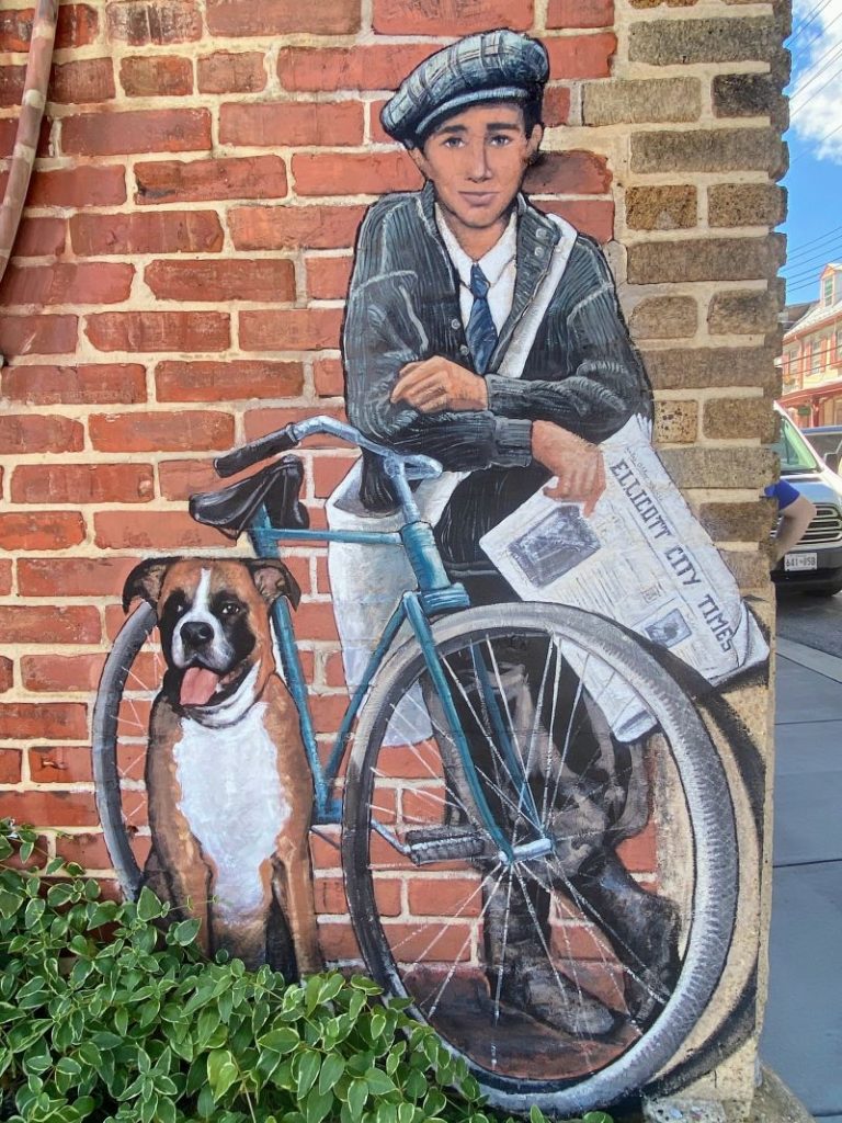 Street art depicting newspaper boy on a bike from the 1940s with his dog by his side.