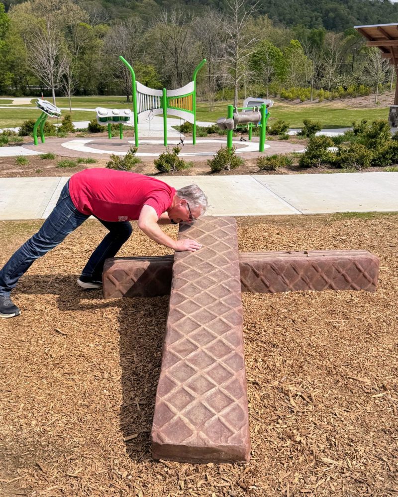 Man pretending to eat giant, fiberglass nutty bar sculpture with musical instrument playground in background on on "Swiss Roll."