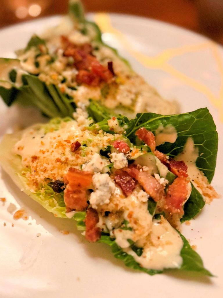 Salad topped with bacon and blue cheese on a white plate trimmed in gold.