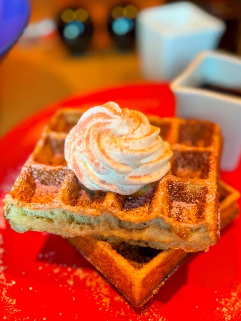 Buckwheat waffles topped with sugar and a swirl of whipped cream sitting on a red plate.