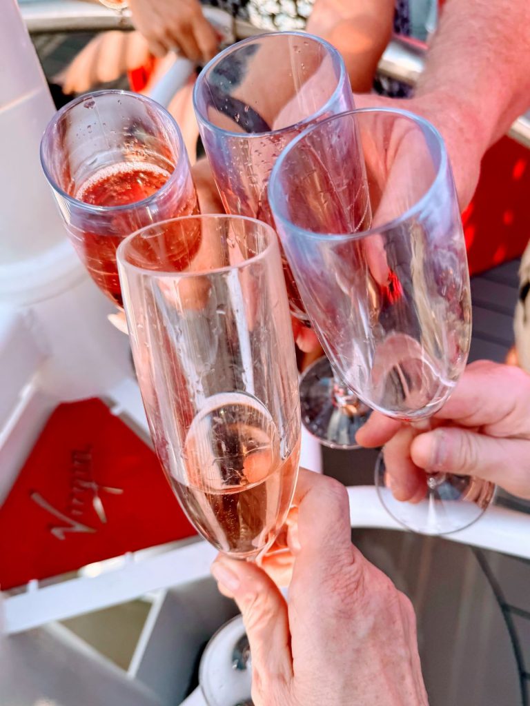 Four champagne glasses clinking to cheer Virgin Voyages food with Virgin logo in background.