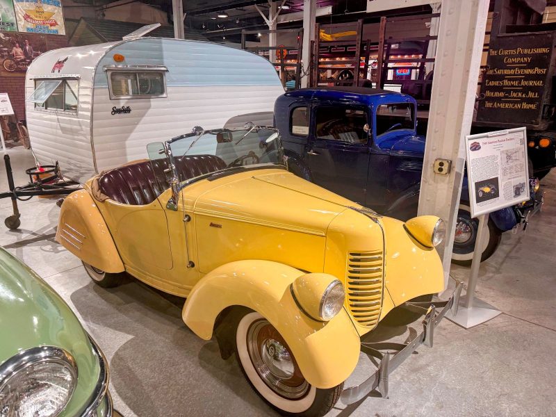 Yellow roadster with blue car and old camper in background.
