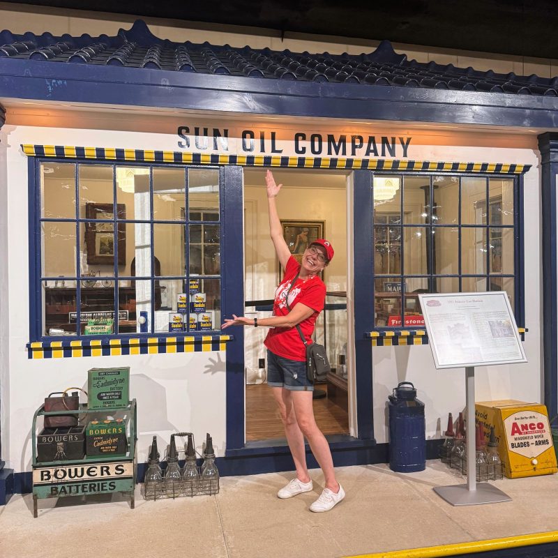 Smiling lady in front of a replica car shop from the early 1900s.
